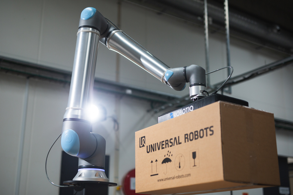 A Universal Robots arm uses a gripper to lift and transfer cardboard boxes in an automated workflow.